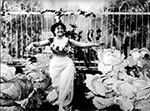 A white woman with long, white skirt and bare arms walks towards camera, surrounded by large cabbages on either side